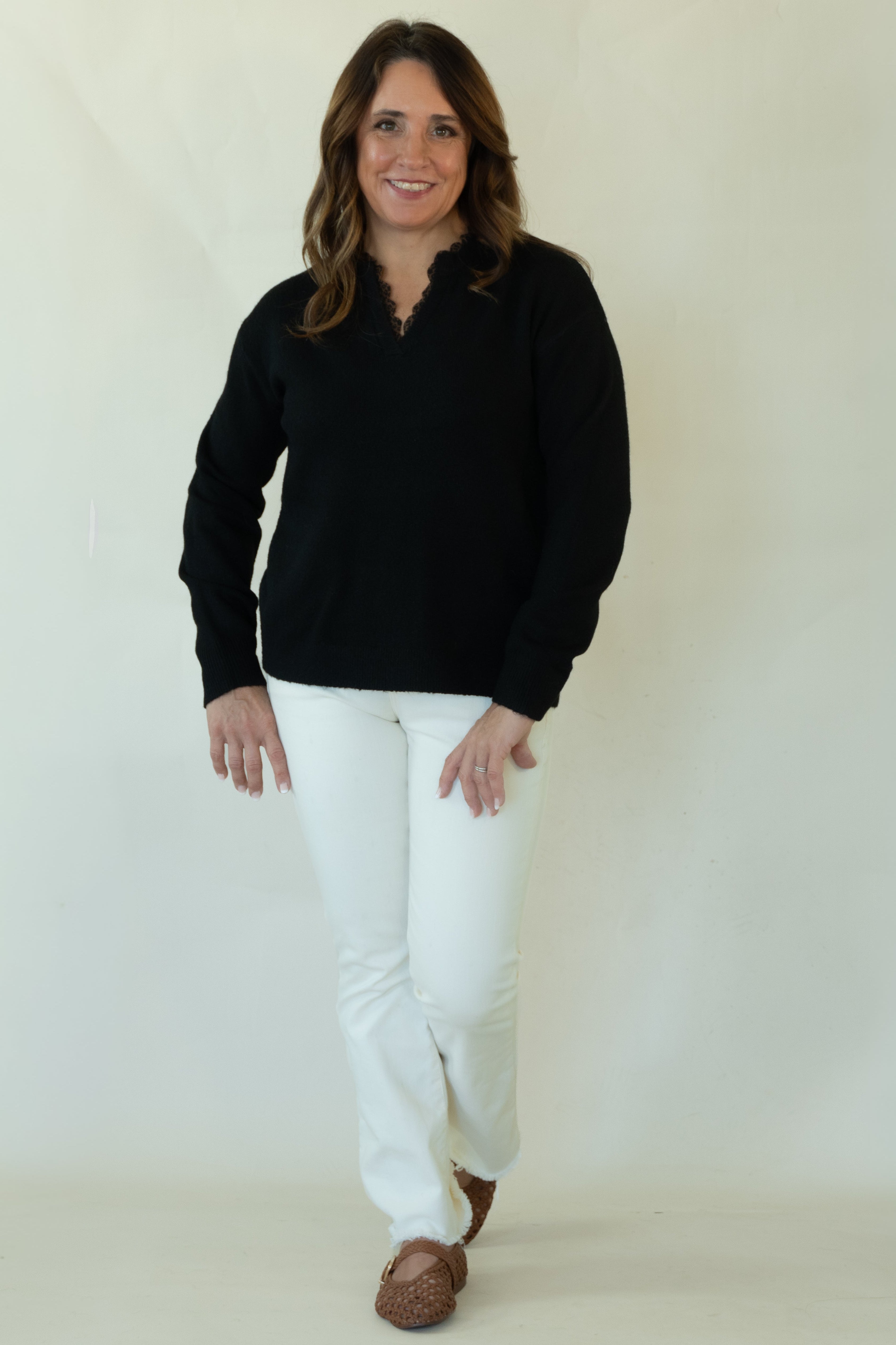 A woman modeling a black long-sleeve knit top with a split neckline, paired with white pants and brown shoes, standing against a plain off-white studio backdrop.