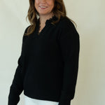 A woman modeling a black long-sleeve knit polo-neck sweater, white pants, and brown woven loafers, standing against a plain off-white studio backdrop.