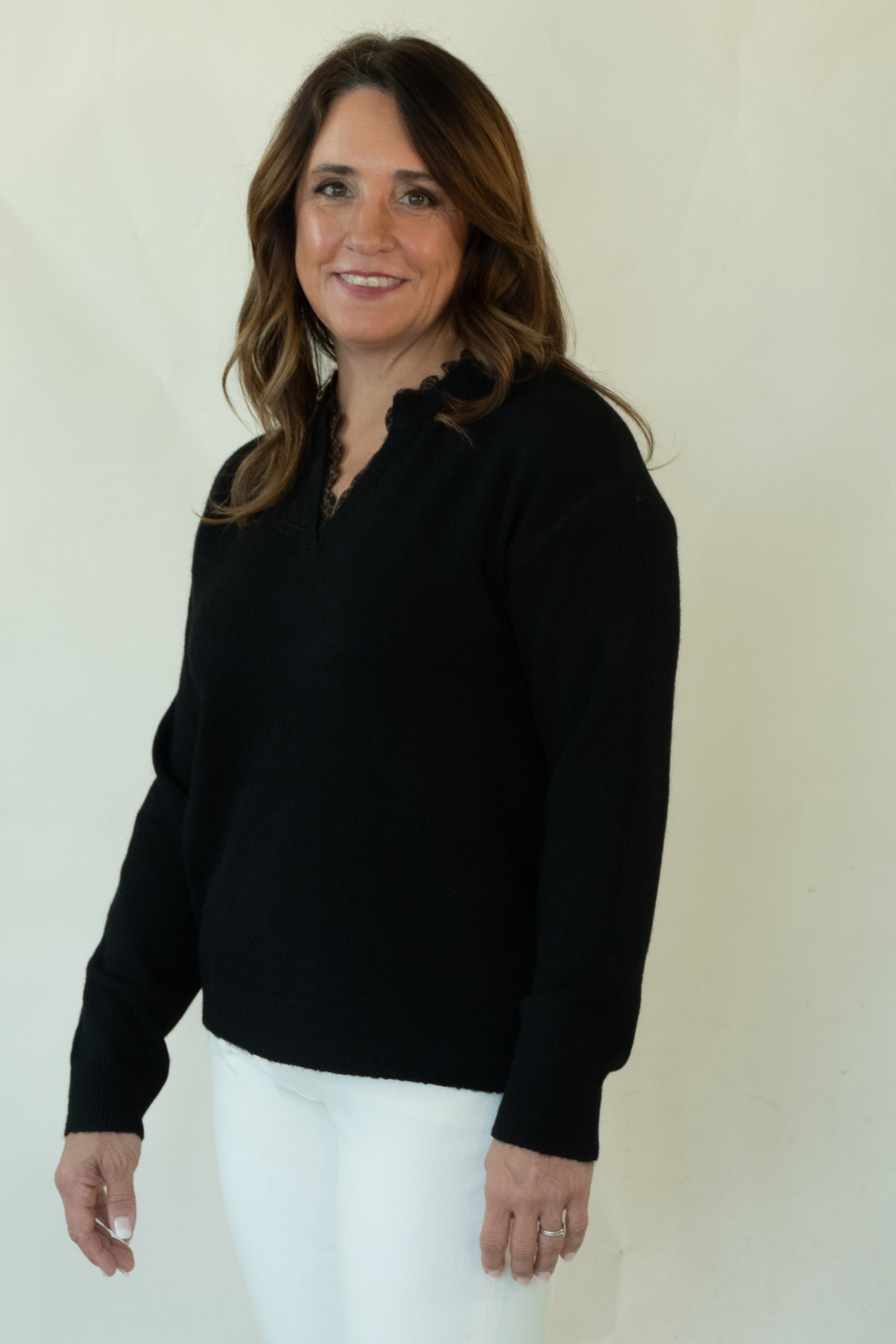 A woman modeling a black long-sleeve knit polo-neck sweater, white pants, and brown woven loafers, standing against a plain off-white studio backdrop.