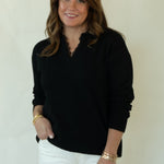 A woman modeling a black long-sleeve knit top and white pants against a plain off-white studio background.