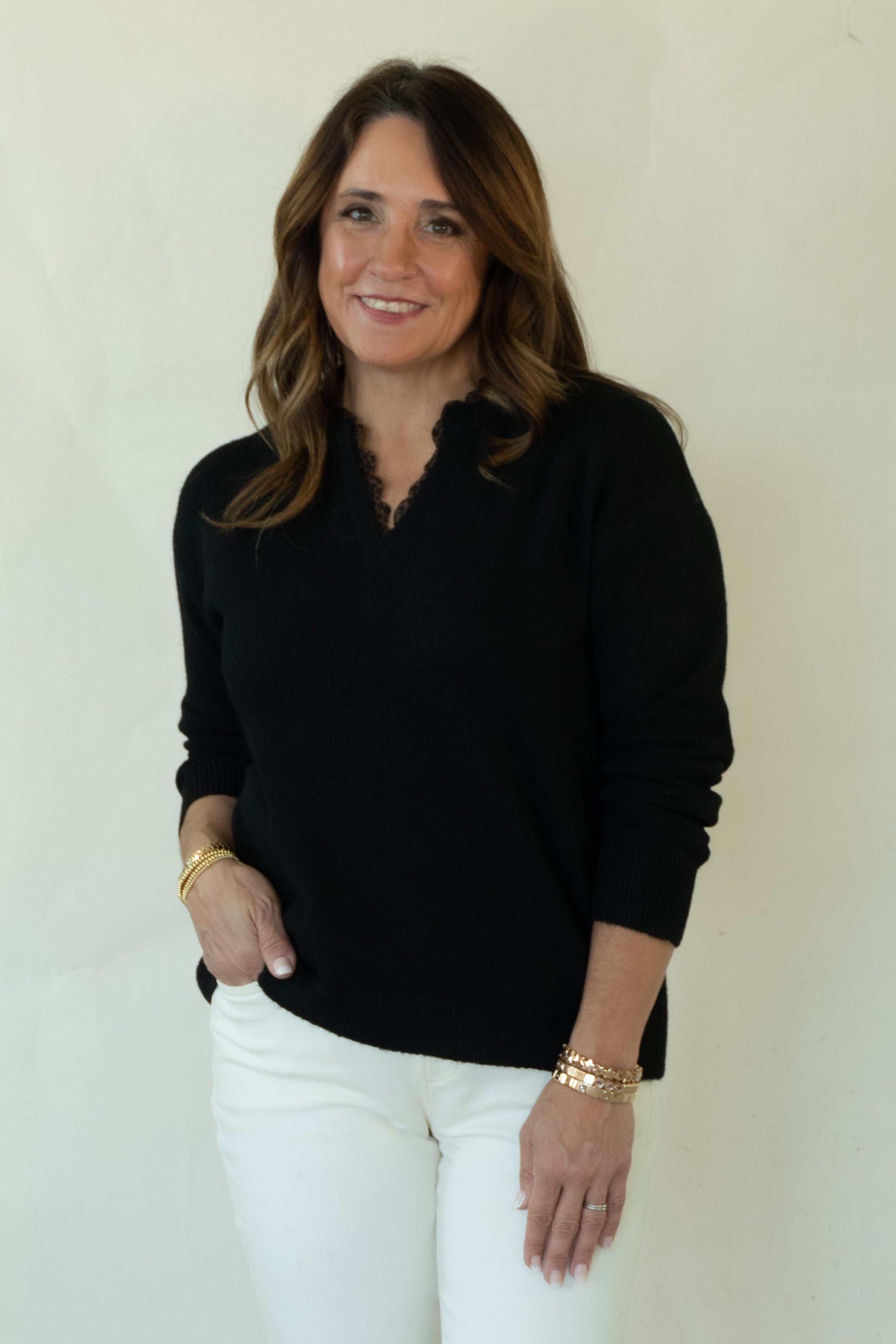 A woman modeling a black long-sleeve knit top and white pants against a plain off-white studio background.