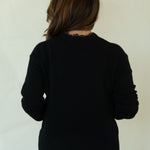 A woman is shown from behind wearing a black long-sleeve top, white pants, and brown shoes, standing against a plain off-white studio background.
