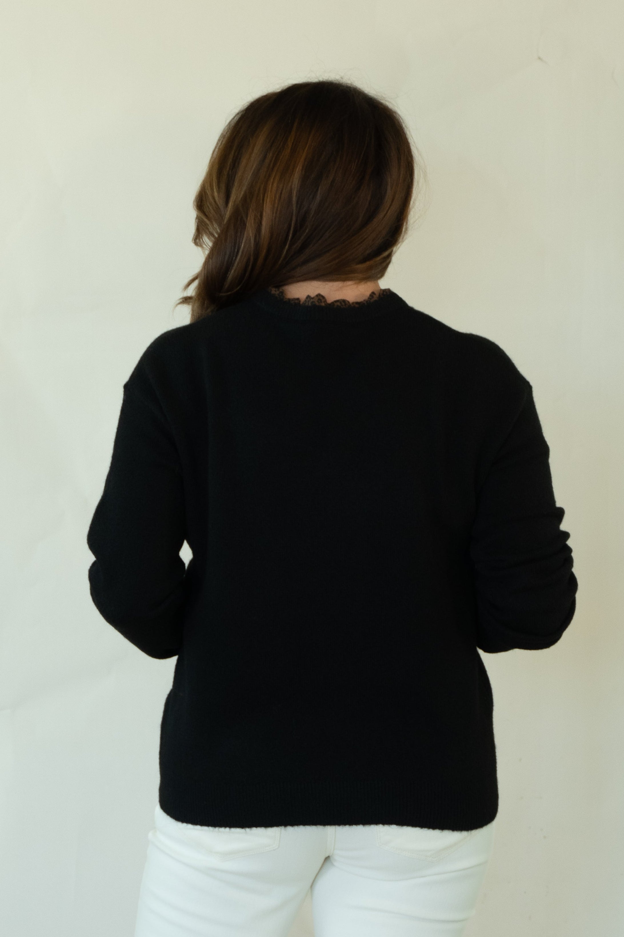 A woman is shown from behind wearing a black long-sleeve top, white pants, and brown shoes, standing against a plain off-white studio background.