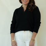 A woman modeling a black long-sleeve mock neck sweater, white pants, and brown woven loafers, standing against a plain off-white studio background.