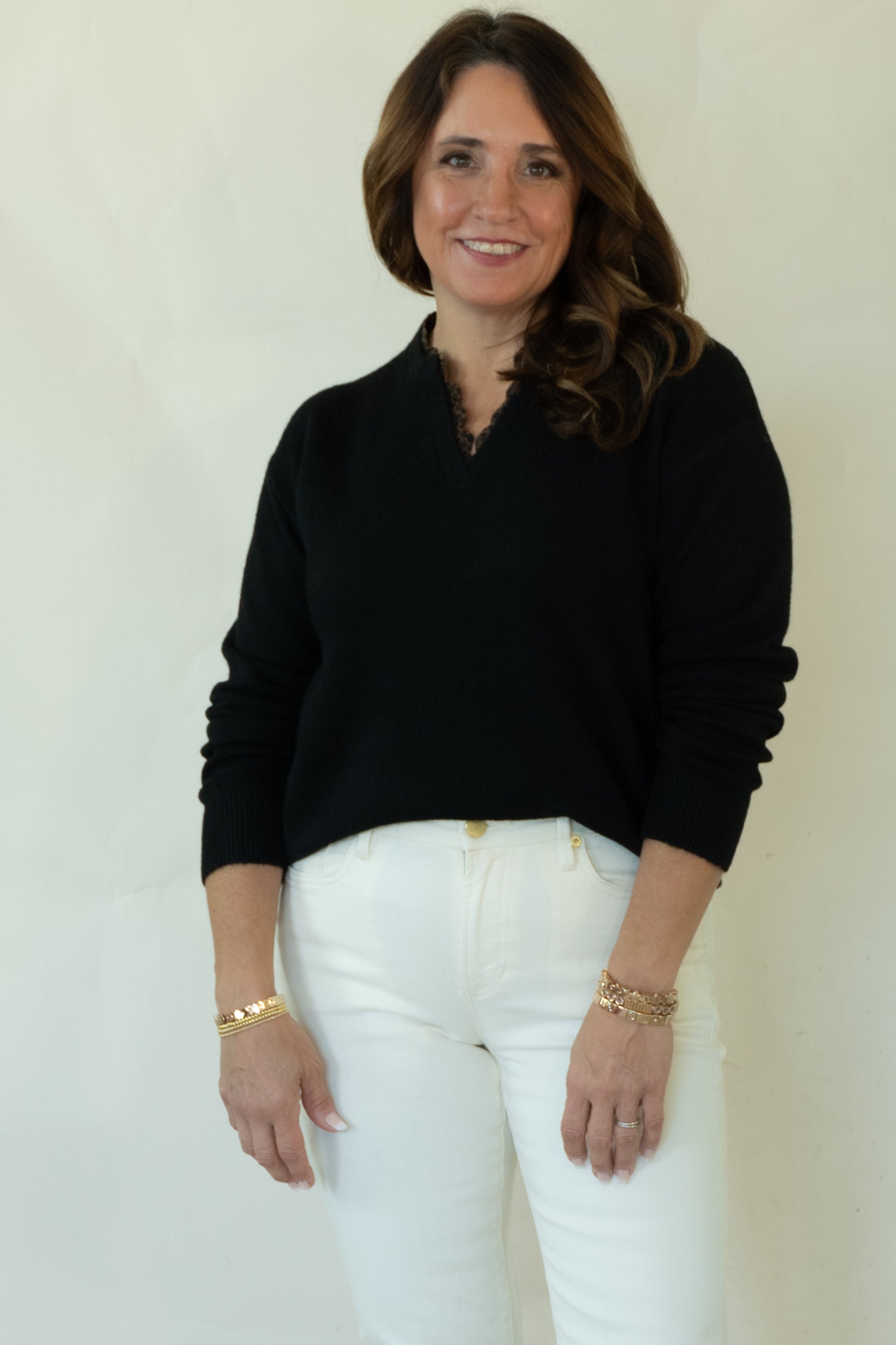 A woman modeling a black long-sleeve mock neck sweater, white pants, and brown woven loafers, standing against a plain off-white studio background.