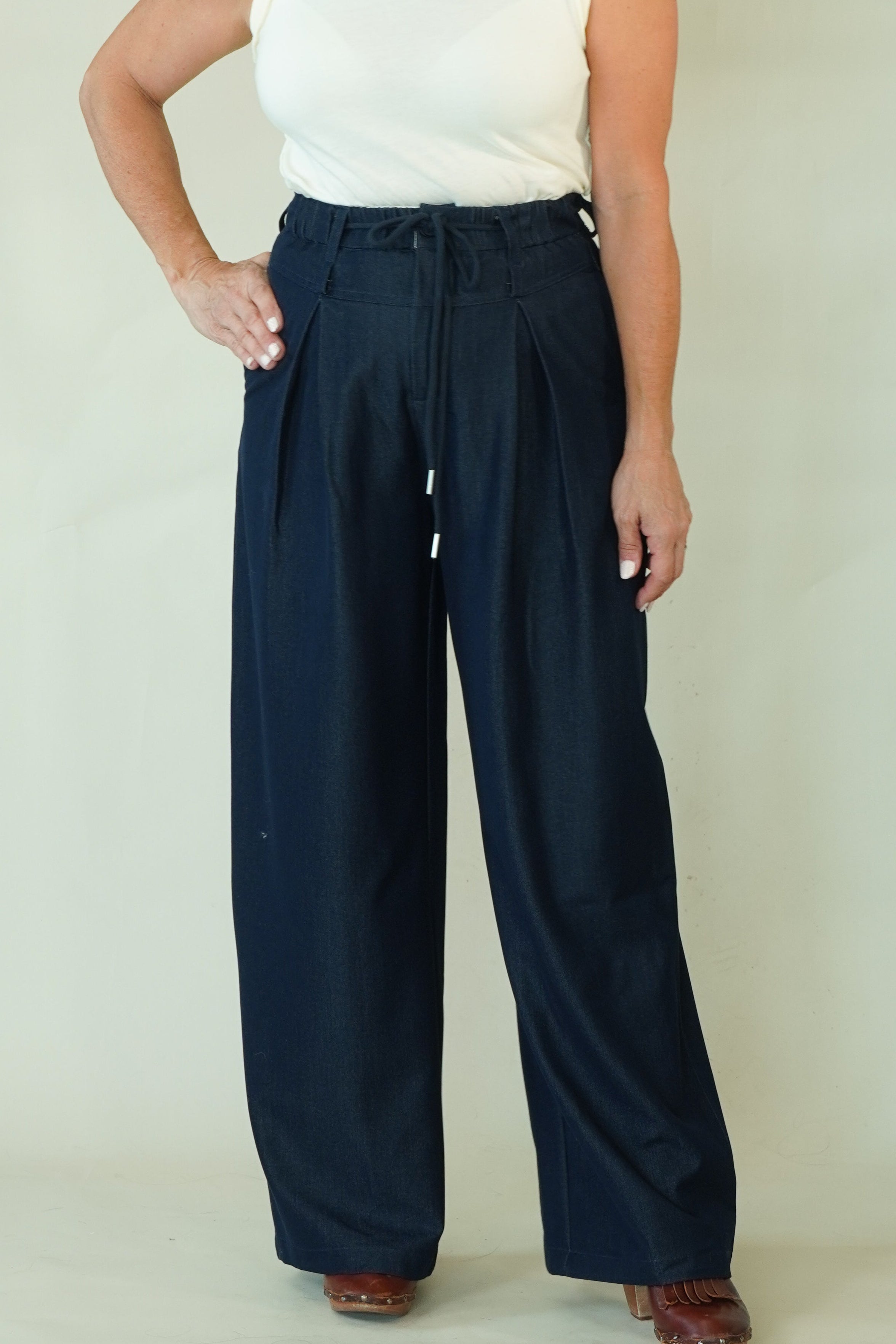 A woman modeling a sleeveless white mock-neck top and dark navy wide-leg trousers with a drawstring waist, standing against a plain light beige studio background.
