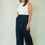 A woman modeling a pair of dark navy wide-leg drawstring pants with a white sleeveless top, standing against a plain light beige studio background.