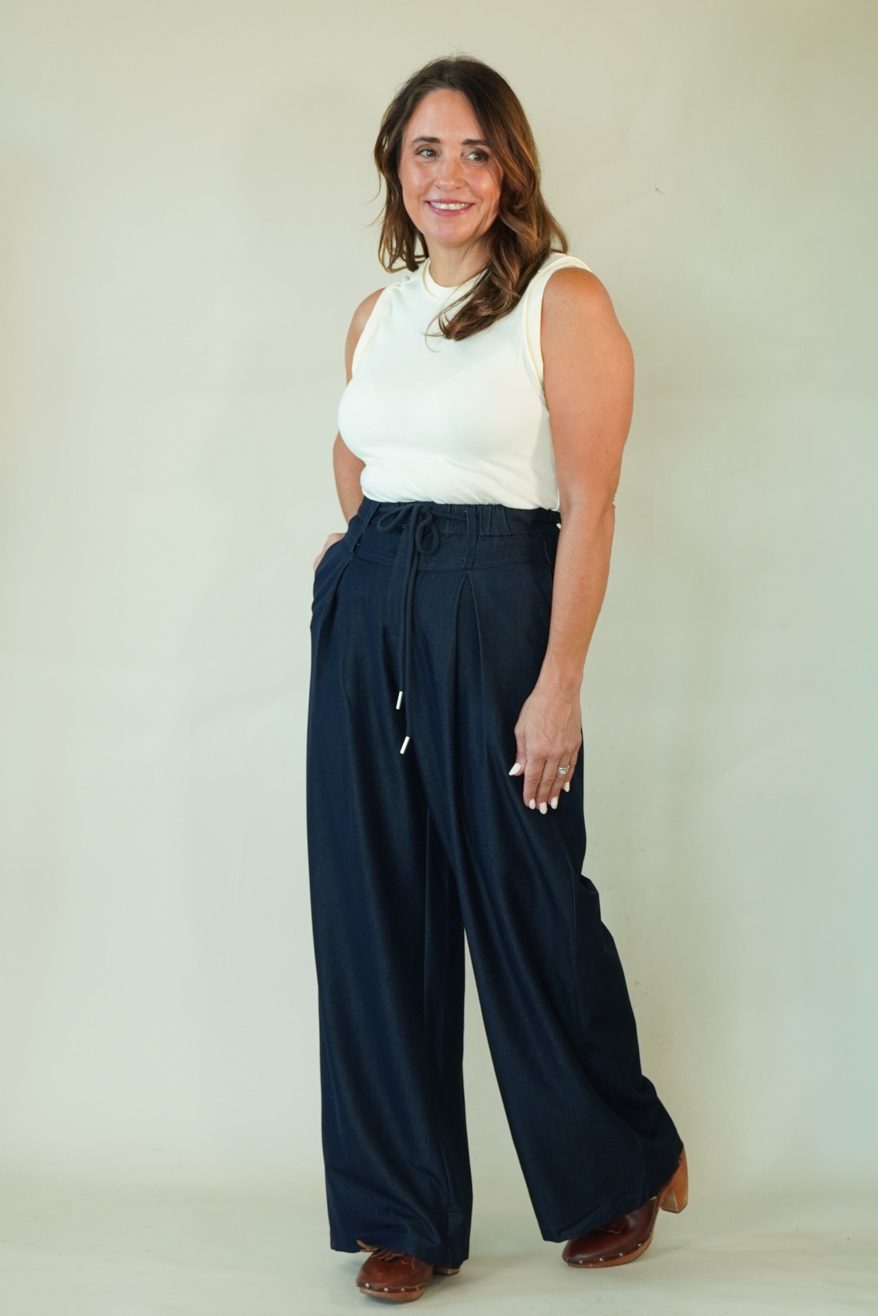 A woman modeling a pair of dark navy wide-leg drawstring pants with a white sleeveless top, standing against a plain light beige studio background.