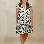 A woman modeling a sleeveless black and white abstract pattern dress with a light blue inner collar and beige sandals, standing against a plain beige studio backdrop.