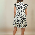 A woman viewed from behind wearing a sleeveless black and white abstract patterned dress with ruffled cap sleeves, paired with beige wedge sandals, against a plain beige studio backdrop.