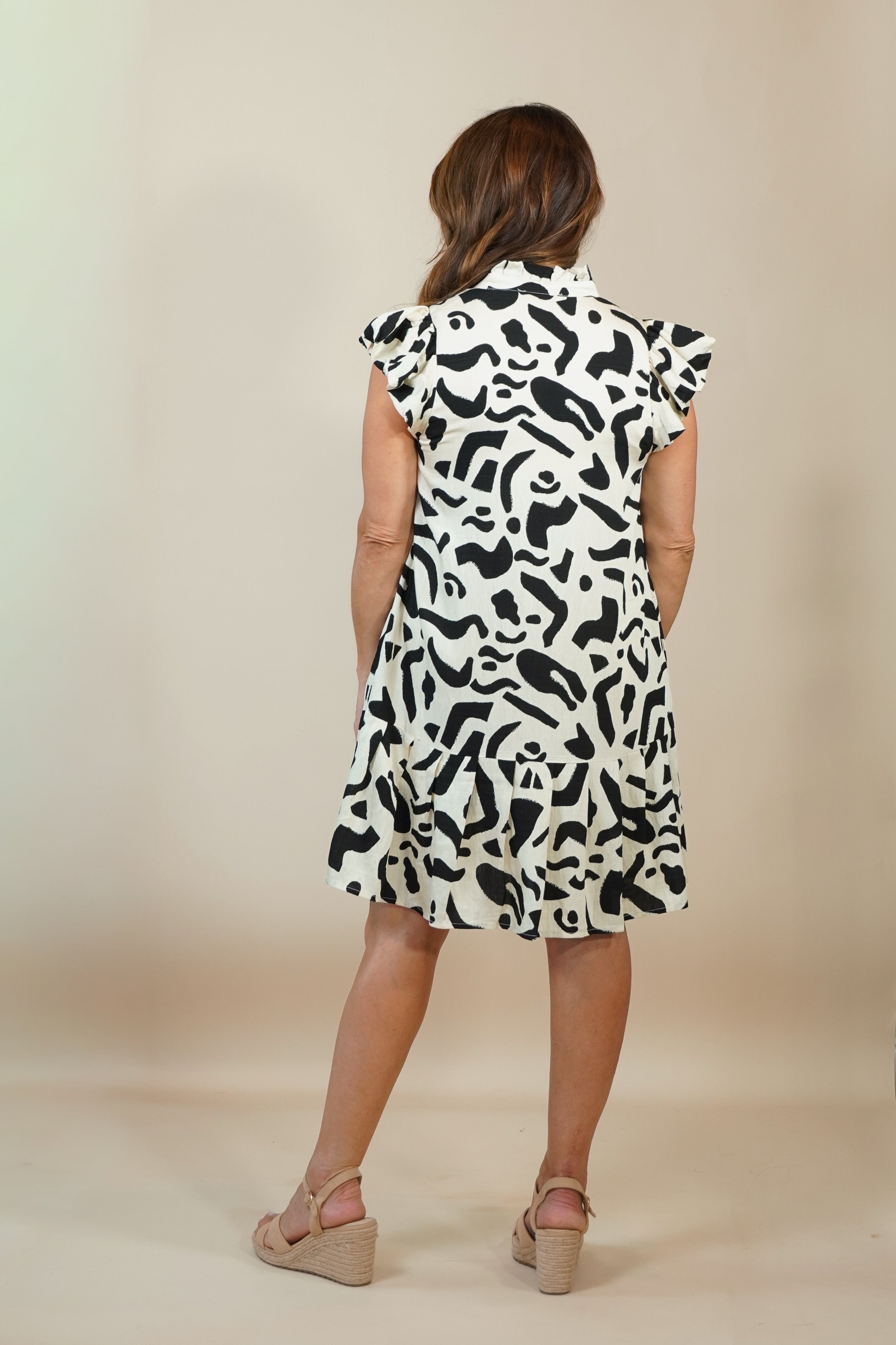 A woman viewed from behind wearing a sleeveless black and white abstract patterned dress with ruffled cap sleeves, paired with beige wedge sandals, against a plain beige studio backdrop.