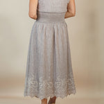 A woman viewed from the back wearing a sleeveless light gray midi dress with a smocked waist and lace hem, paired with tan wedge sandals, against a plain beige studio backdrop.