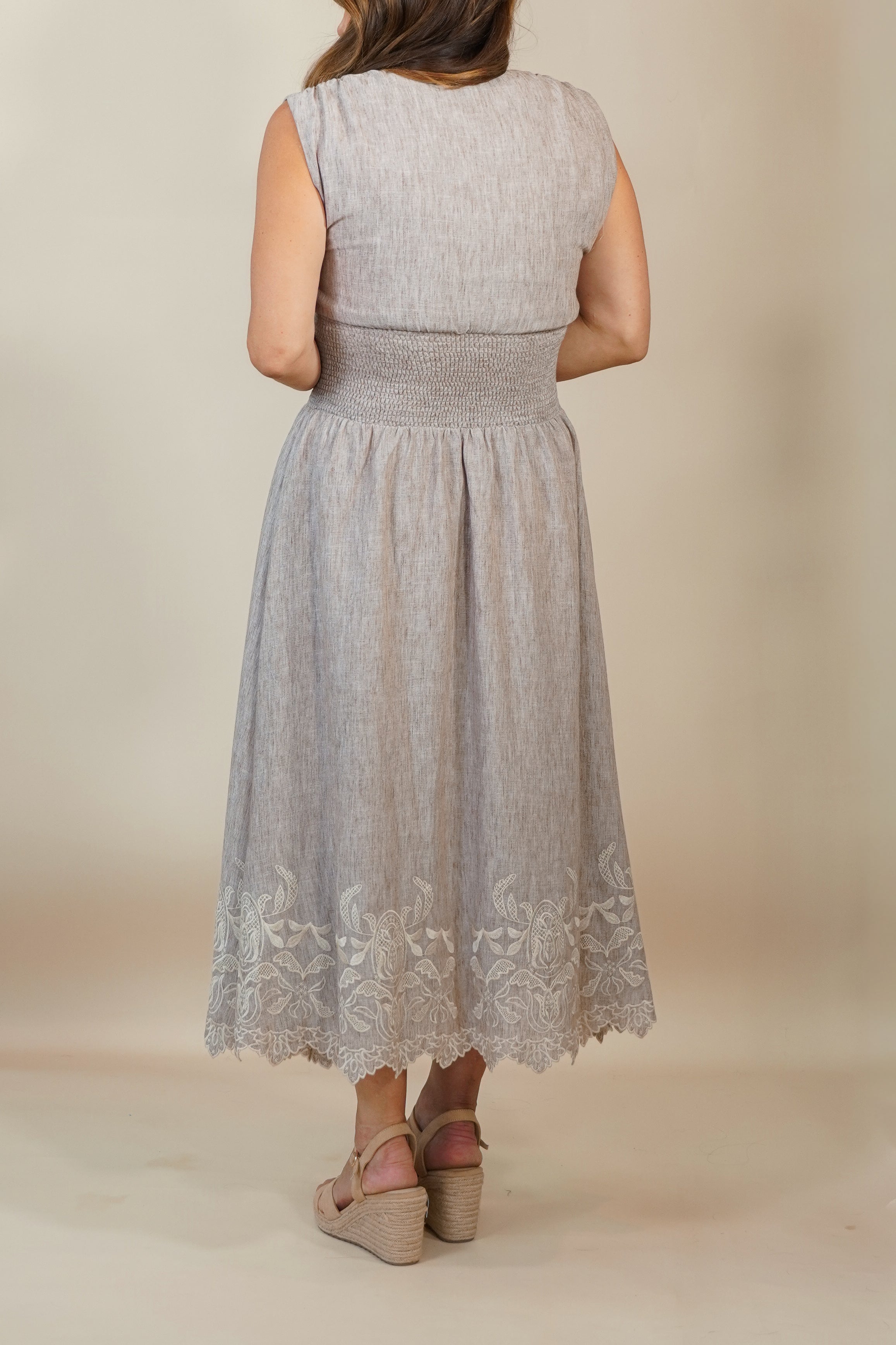 A woman viewed from the back wearing a sleeveless light gray midi dress with a smocked waist and lace hem, paired with tan wedge sandals, against a plain beige studio backdrop.