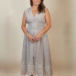 A woman modeling a sleeveless light gray linen midi dress with a V-neckline, buttoned front, and lace-trimmed hem, paired with beige wedge sandals against a neutral beige studio backdrop.