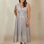 A woman modeling a sleeveless light gray midi dress with a V-neckline, buttoned front, and lace hem, standing against a plain beige studio backdrop.