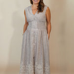 A woman modeling a sleeveless light gray midi dress with a textured pattern, V-neckline, and lace detailing at the hem, paired with beige wedge sandals against a plain beige studio background.