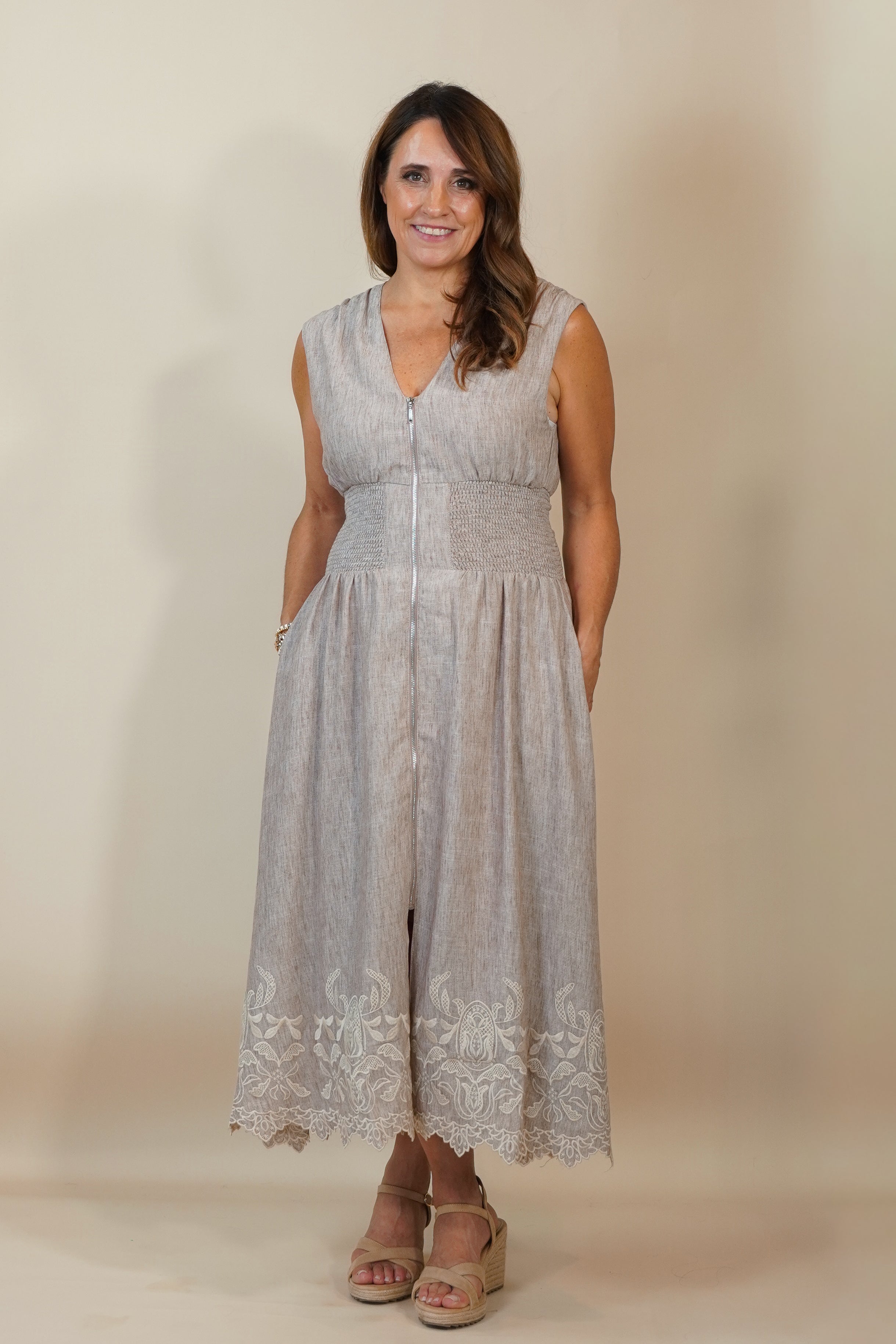 A woman modeling a sleeveless light gray midi dress with a textured pattern, V-neckline, and lace detailing at the hem, paired with beige wedge sandals against a plain beige studio background.