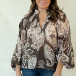 A woman modeling a brown and beige paisley patterned blouse with blue jeans and tan boots, standing against a plain white studio background.
