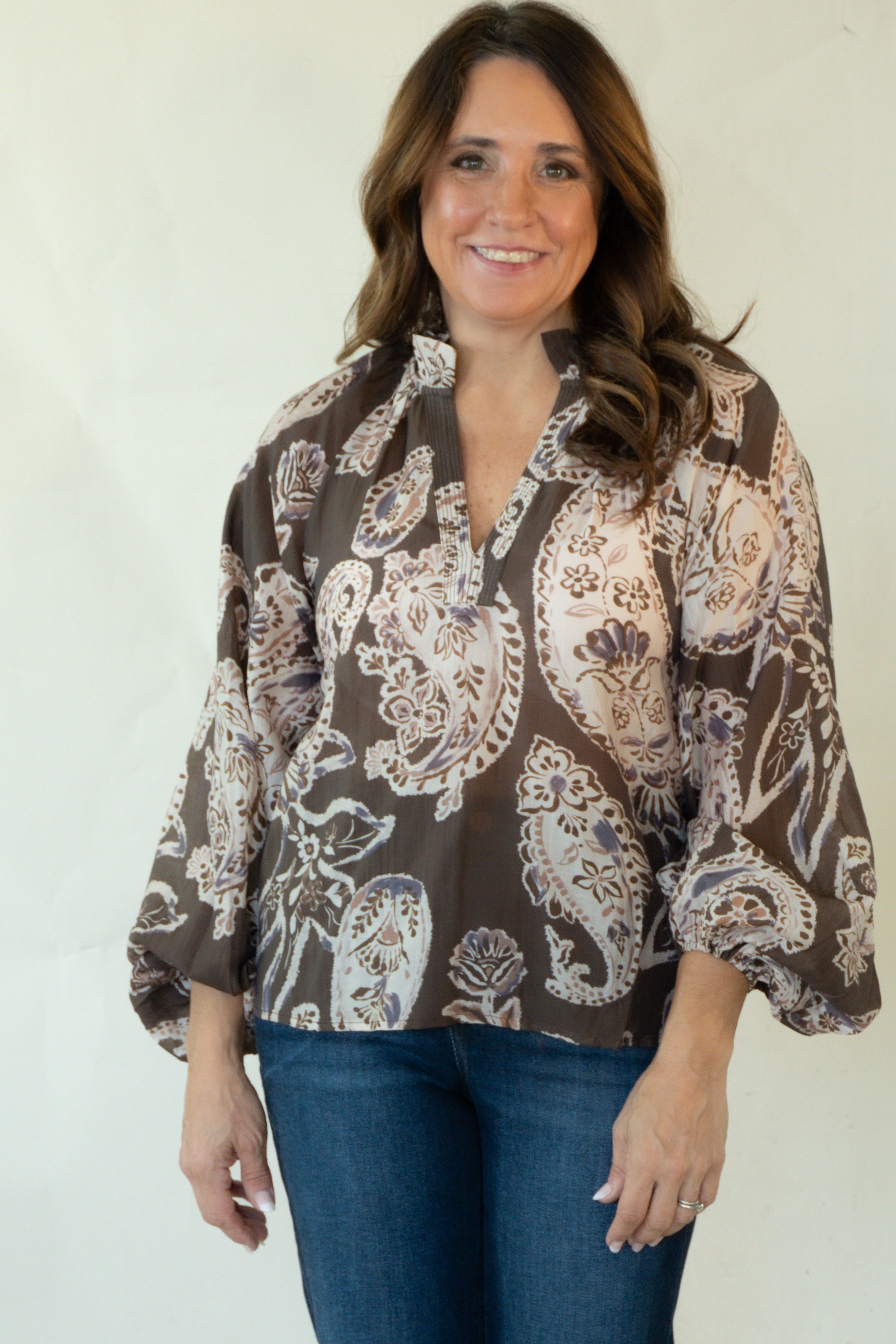 A woman modeling a brown and beige paisley patterned blouse with blue jeans and tan boots, standing against a plain white studio background.