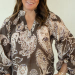A woman modeling a brown and beige paisley patterned blouse with white pants and brown woven flats, standing against a plain off-white studio background.