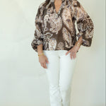 A woman modeling a brown and beige paisley print blouse with white pants and brown woven shoes, standing against a plain off-white studio background.