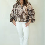 A woman modeling a brown and beige paisley patterned blouse with white pants and brown woven shoes, standing against a plain off-white studio backdrop.