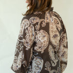 A woman is shown from the back wearing a brown long-sleeve button-up shirt with a beige paisley pattern, blue jeans, and brown loafers, standing against a plain off-white studio background.