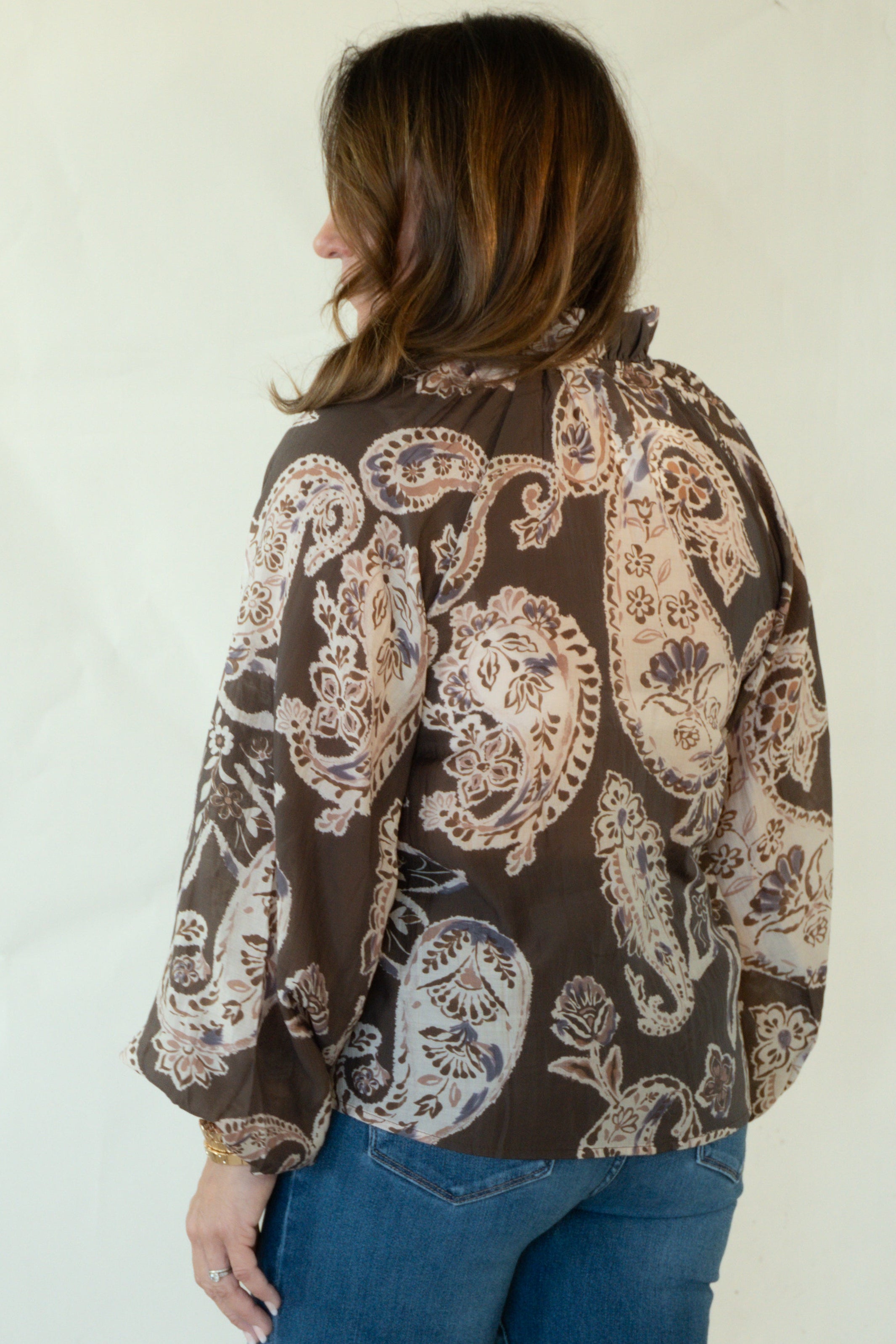 A woman is shown from the back wearing a brown long-sleeve button-up shirt with a beige paisley pattern, blue jeans, and brown loafers, standing against a plain off-white studio background.