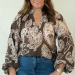 A woman modeling a brown paisley print button-down shirt with blue jeans and brown shoes, standing against a white studio backdrop.