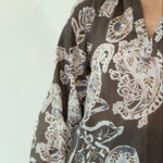 A woman modeling a brown long-sleeve button-up blouse with a beige paisley pattern, paired with dark blue jeans and tan boots, standing against a plain white studio backdrop.