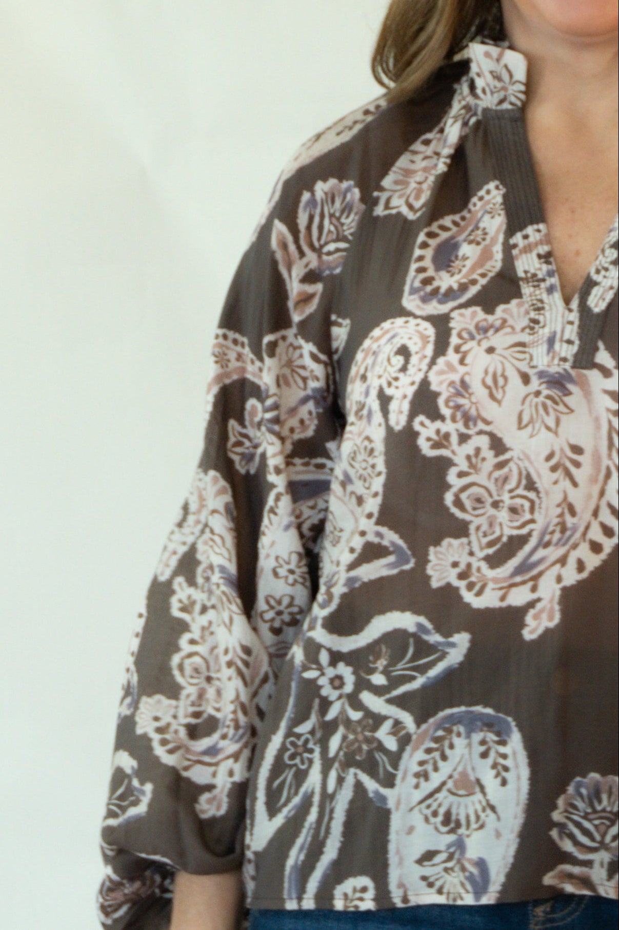 A woman modeling a brown long-sleeve button-up blouse with a beige paisley pattern, paired with dark blue jeans and tan boots, standing against a plain white studio backdrop.