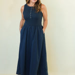 A woman modeling a sleeveless navy blue maxi dress with a buttoned neckline and side pockets, paired with beige wedge sandals, against a plain cream studio backdrop.