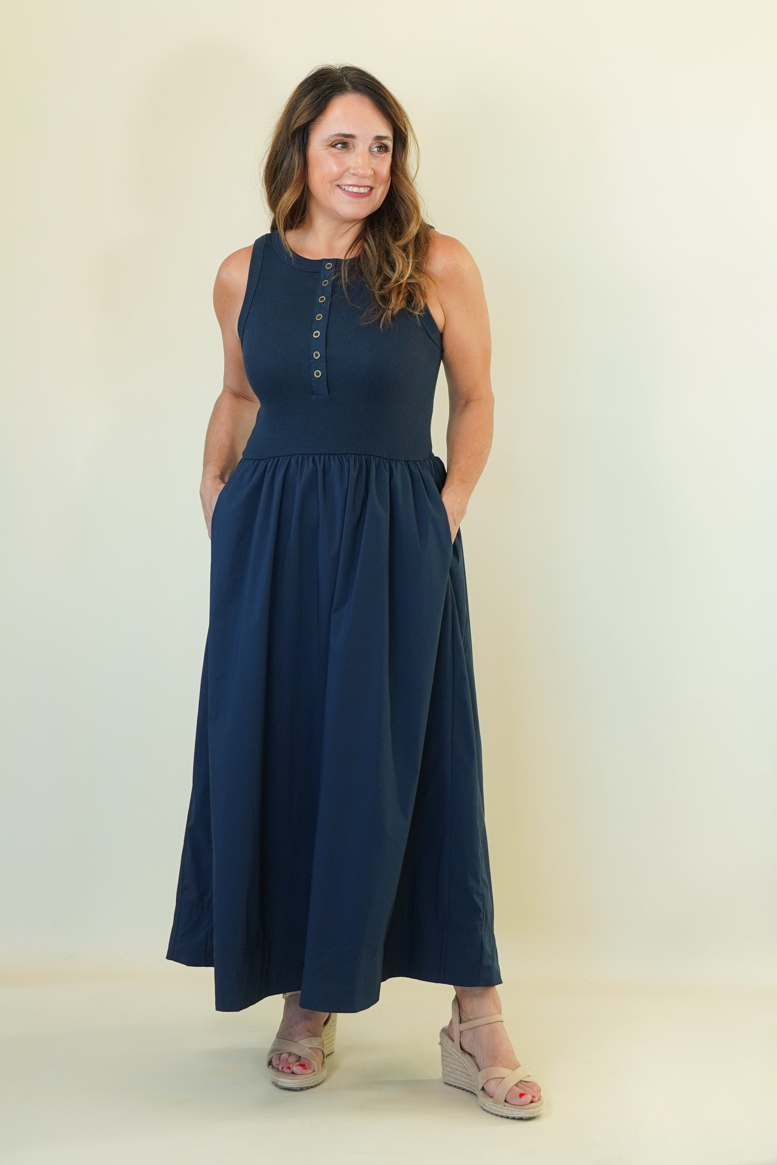 A woman modeling a sleeveless navy blue maxi dress with a buttoned neckline and side pockets, paired with beige wedge sandals, against a plain cream studio backdrop.