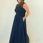 A woman modeling a sleeveless navy blue maxi dress with a buttoned neckline and side pockets, standing against a plain off-white studio background.