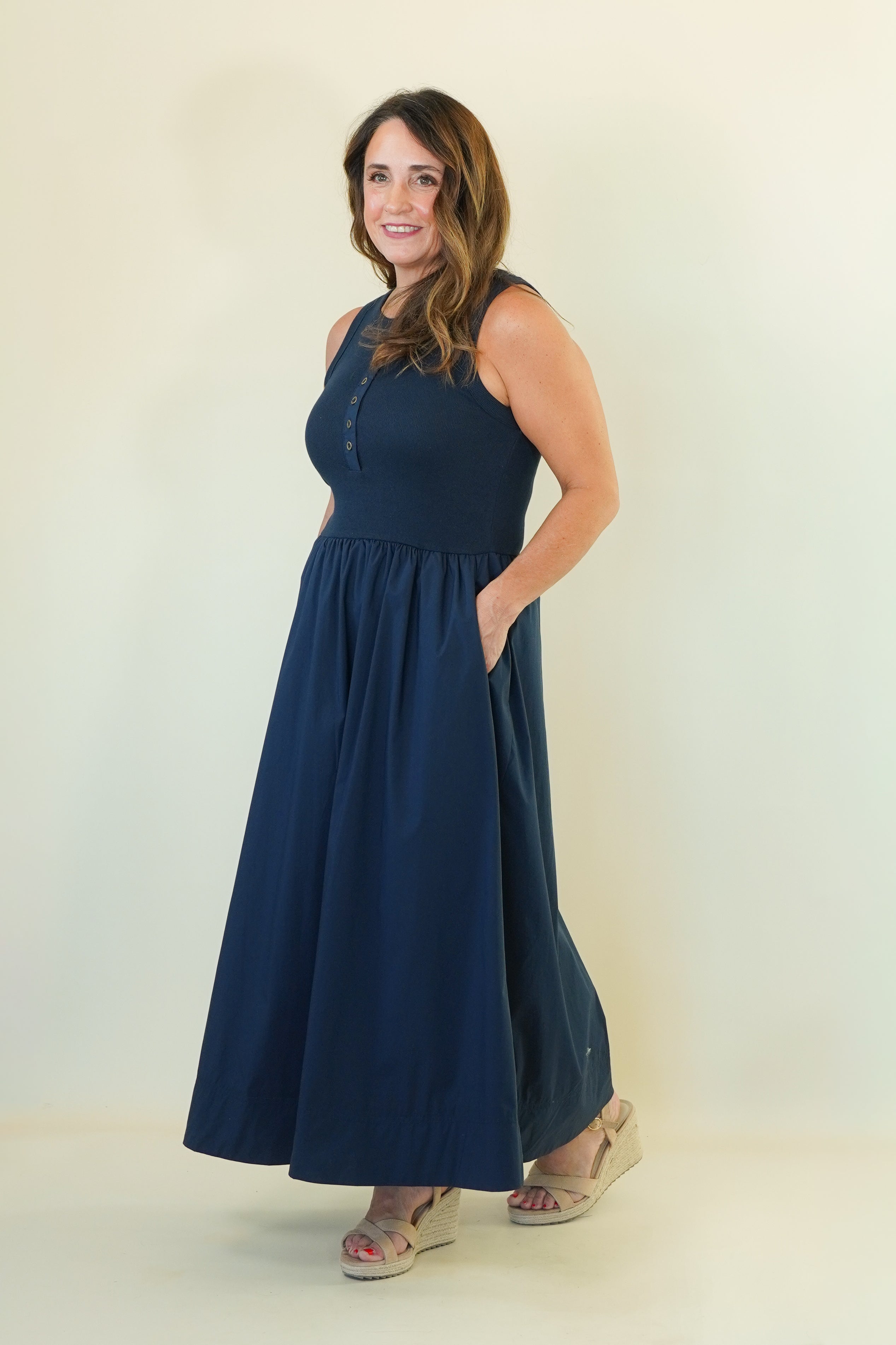 A woman modeling a sleeveless navy blue maxi dress with a buttoned neckline and side pockets, standing against a plain off-white studio background.