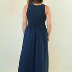 A woman viewed from behind wearing a sleeveless navy blue maxi dress with a gathered waist, standing against a plain off-white studio background.