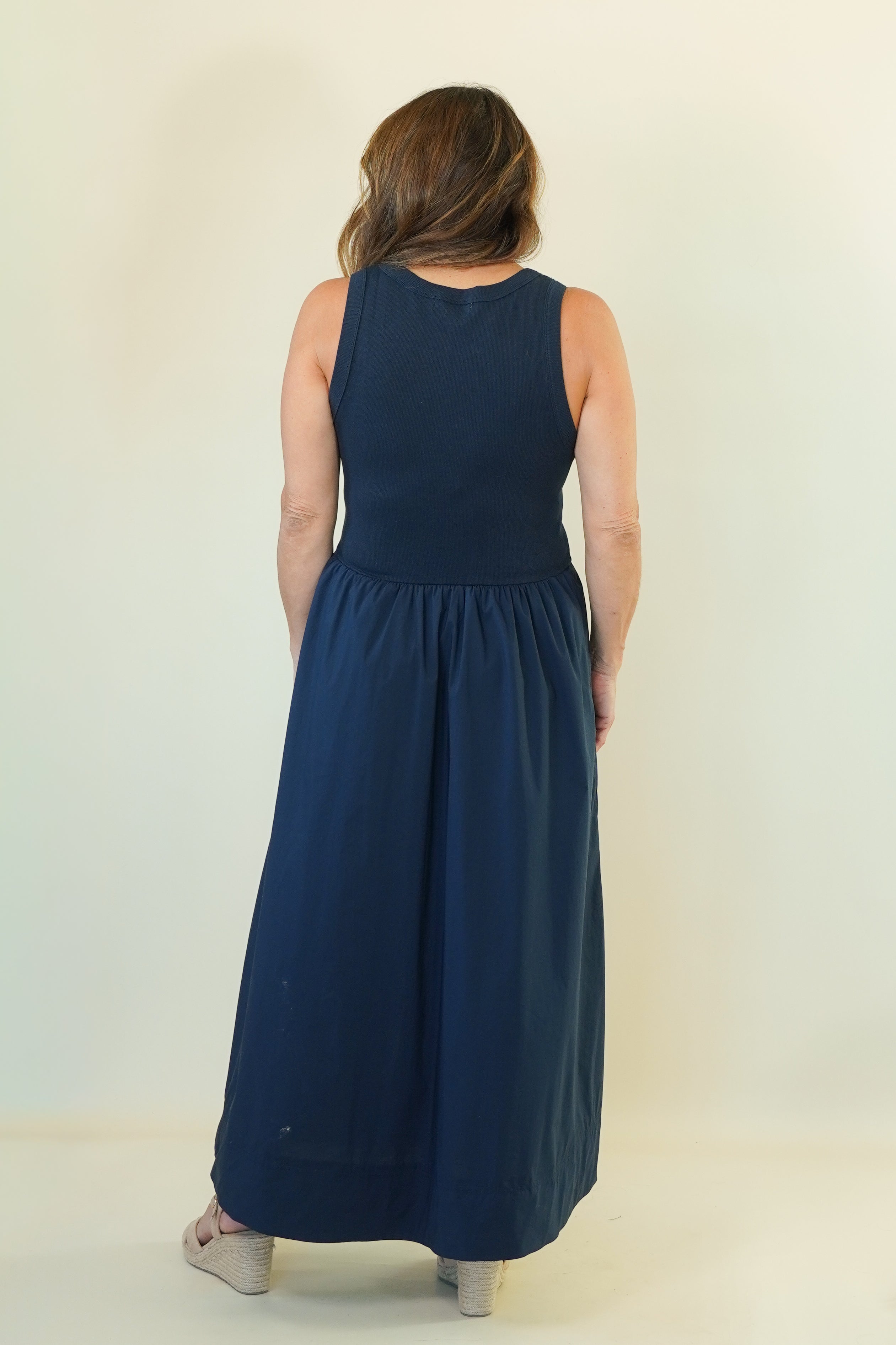 A woman viewed from behind wearing a sleeveless navy blue maxi dress with a gathered waist, standing against a plain off-white studio background.