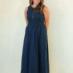 A woman modeling a sleeveless navy blue maxi dress with a buttoned neckline and a gathered waist, standing against a plain off-white studio background.