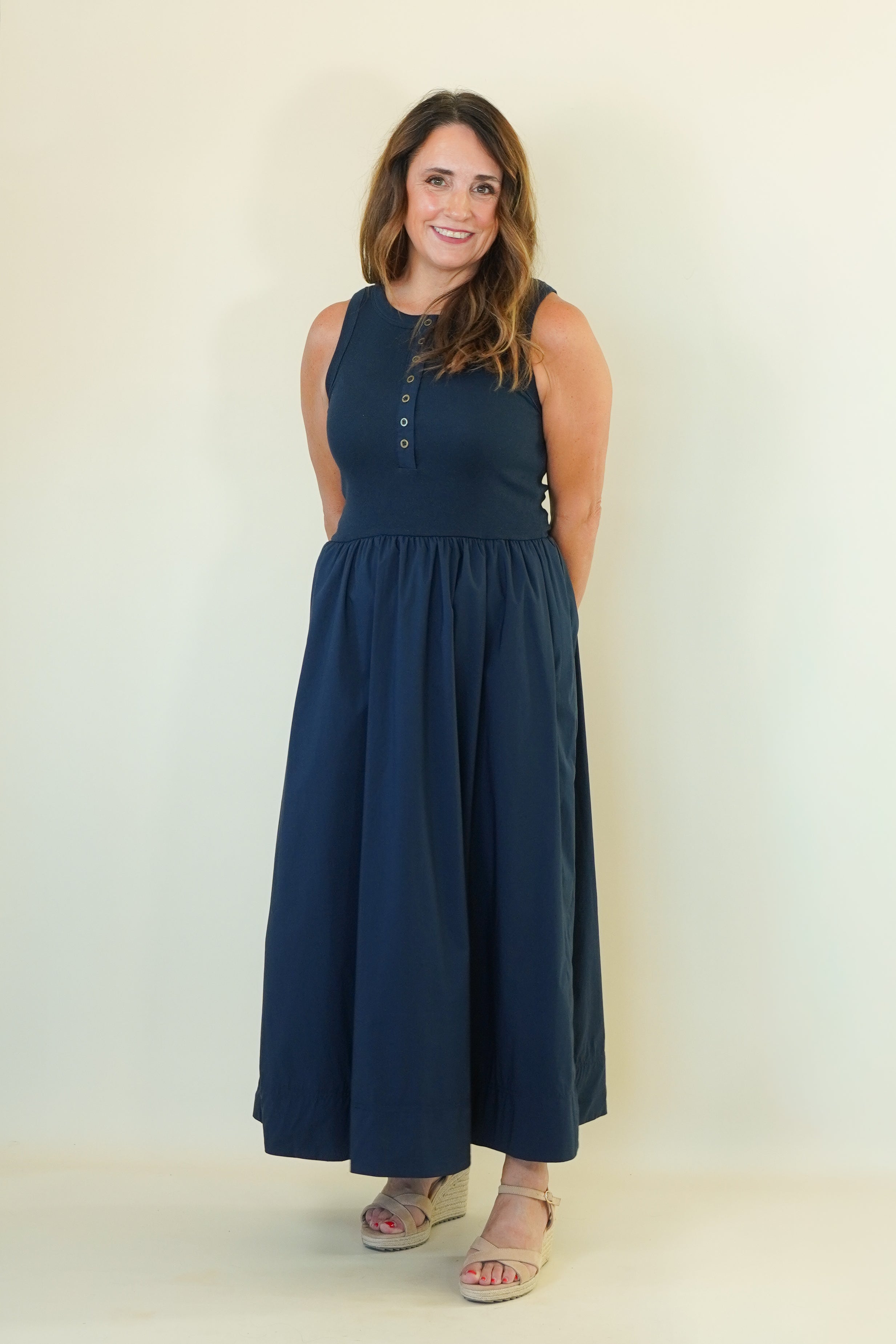 A woman modeling a sleeveless navy blue maxi dress with a buttoned neckline and a gathered waist, standing against a plain off-white studio background.
