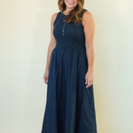 A woman modeling a sleeveless navy blue maxi dress with a buttoned neckline, standing against a plain cream-colored studio background.