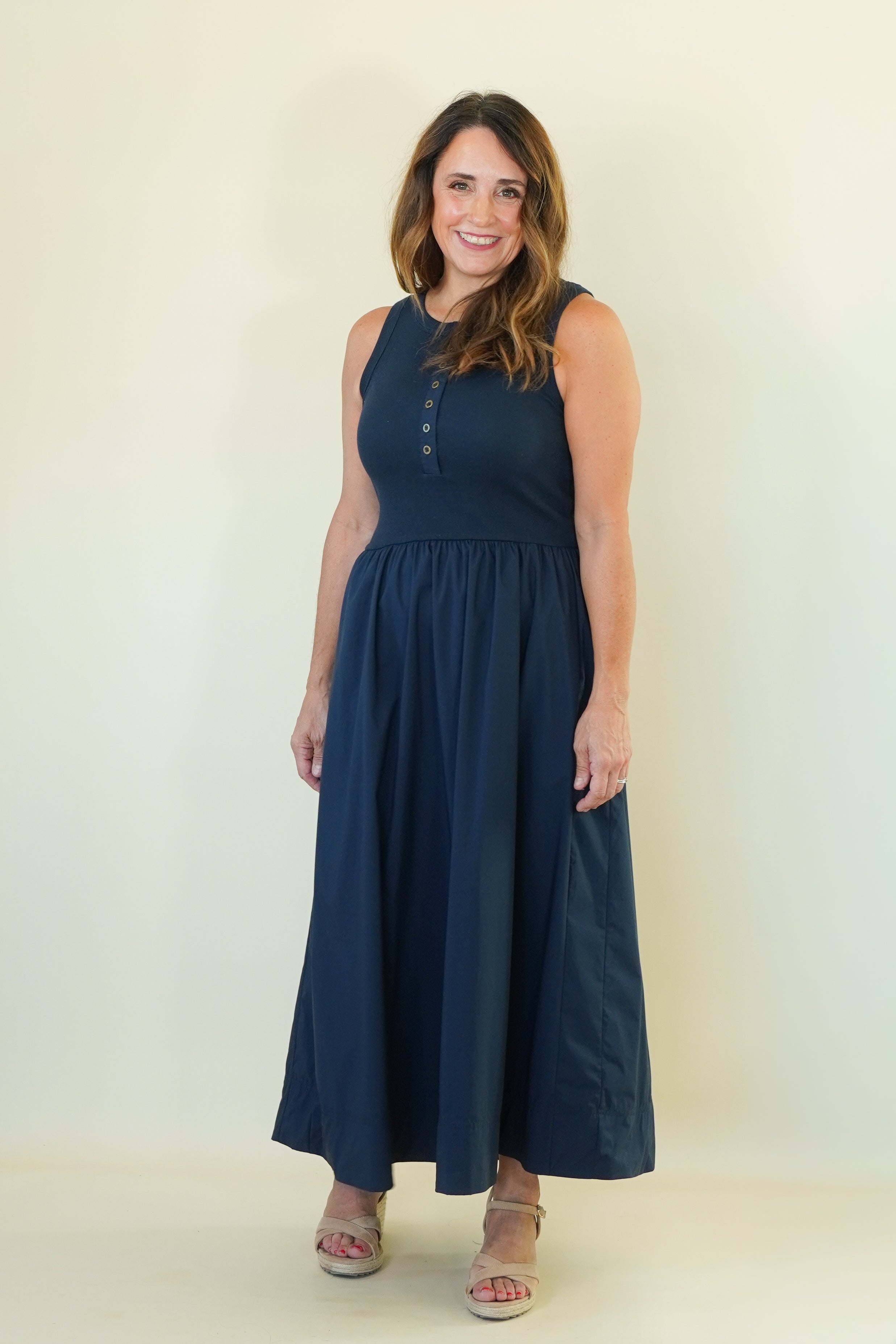 A woman modeling a sleeveless navy blue maxi dress with a buttoned neckline, standing against a plain cream-colored studio background.