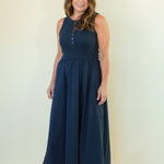 A woman modeling a sleeveless navy blue maxi dress with a buttoned neckline and a gathered waist, standing against a plain off-white studio background.