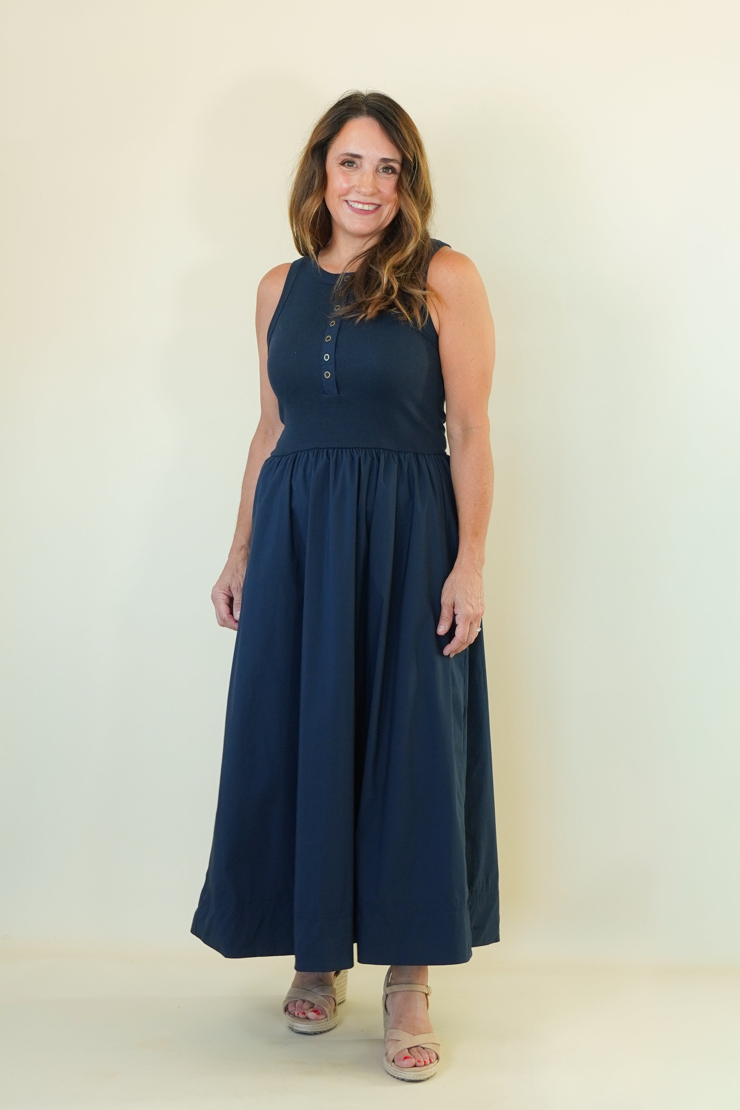 A woman modeling a sleeveless navy blue maxi dress with a buttoned neckline and a gathered waist, standing against a plain off-white studio background.
