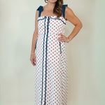A woman modeling a white sleeveless maxi dress with red polka dots and dark blue trim, including shoulder straps and a front tie closure, against a plain light beige studio background.