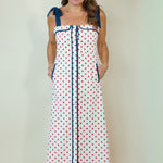 A woman modeling a white sleeveless maxi dress with red polka dots, navy blue trim, and tie shoulder straps, standing against a plain light beige background.