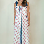 A woman modeling a white sleeveless maxi dress with red polka dots, blue shoulder ties, and a blue vertical trim detail, set against a plain cream background.