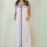 A woman modeling a white sleeveless maxi dress with red polka dots and blue trim details, including shoulder ties and a front button placket, set against a plain light beige studio background.