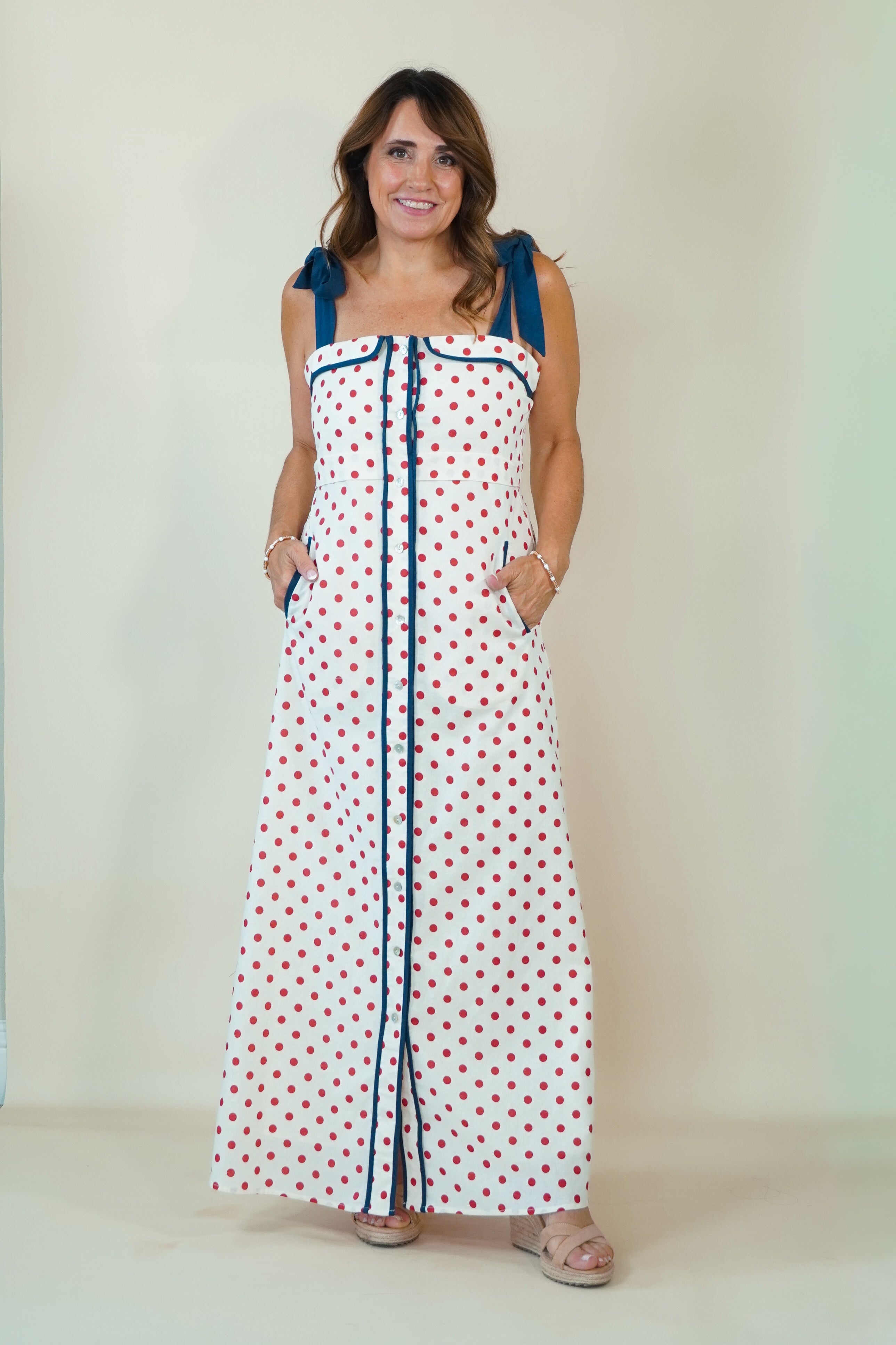 A woman modeling a white sleeveless maxi dress with red polka dots and blue trim details, including shoulder ties and a front button placket, set against a plain light beige studio background.