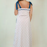 A woman viewed from behind wearing a white sleeveless maxi dress with red polka dots and blue shoulder ties, standing against a plain off-white studio background.