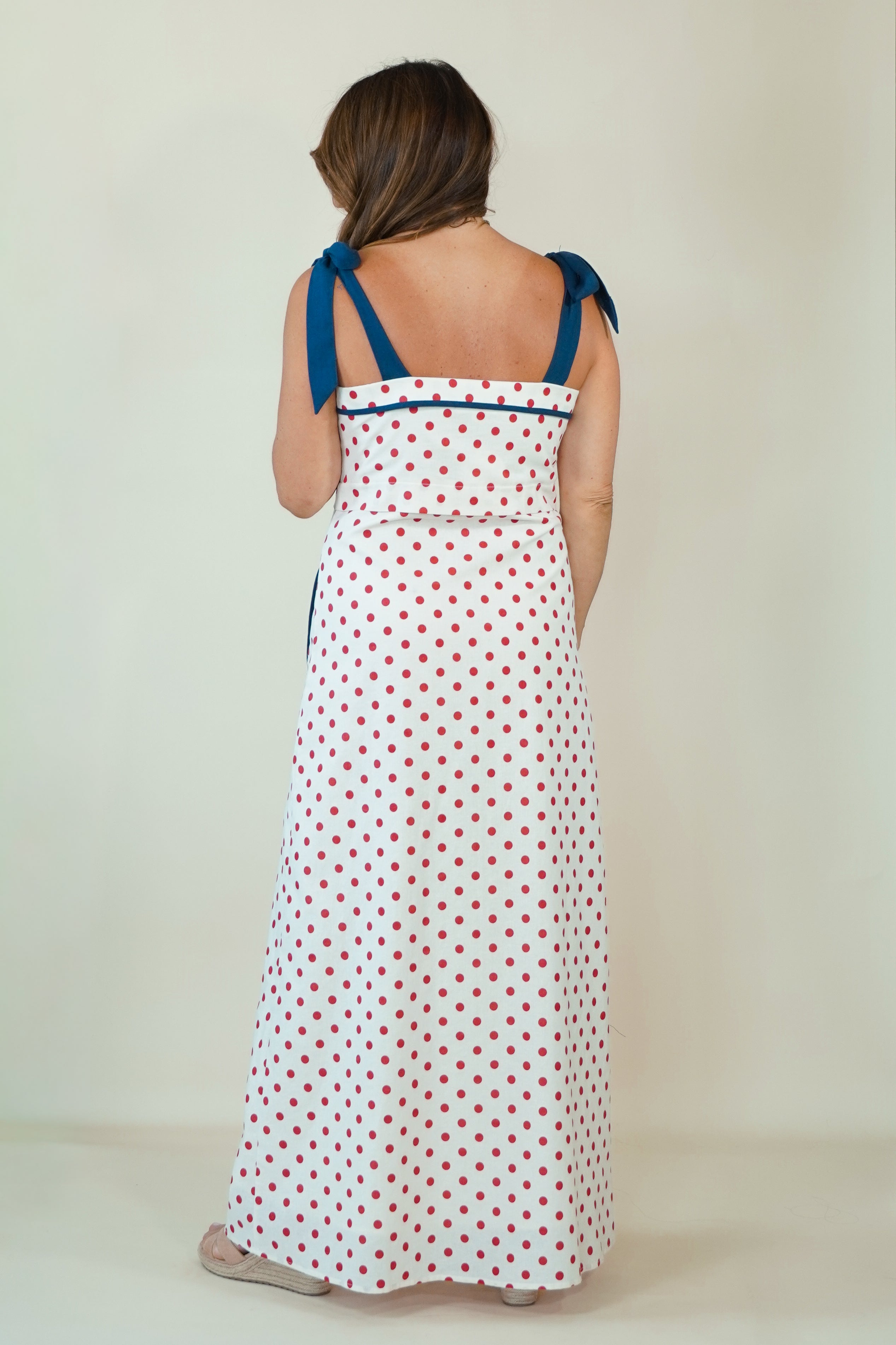 A woman viewed from behind wearing a white sleeveless maxi dress with red polka dots and blue shoulder ties, standing against a plain off-white studio background.